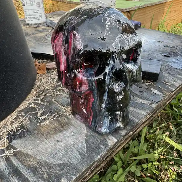 A glossy, partially painted skull sculpture with black, red, and purple paint dripping down its left side sits on a…