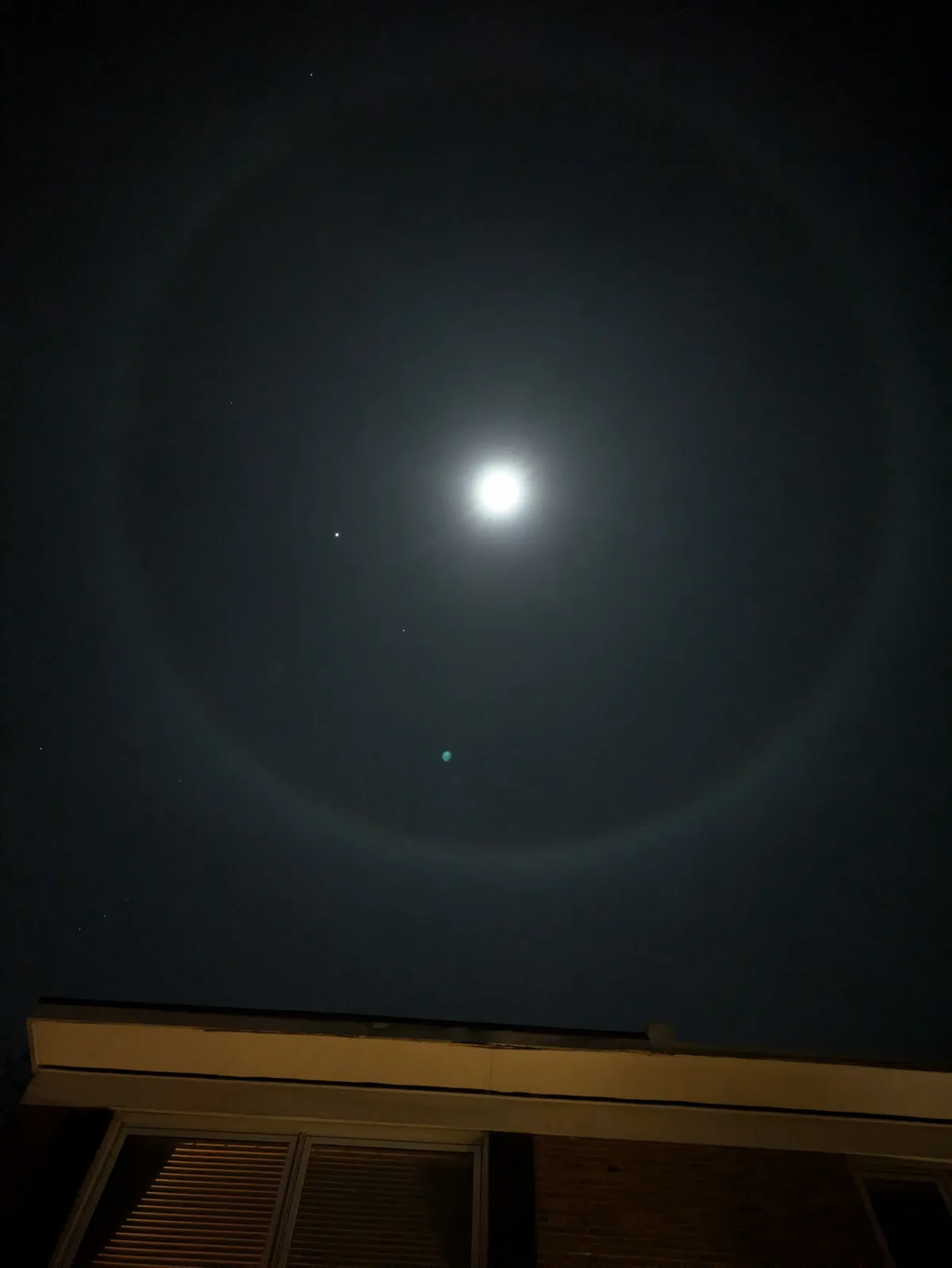 A night sky photo shows a bright moon surrounded by a faint, circular halo of light, with a few stars visible nearby and the…