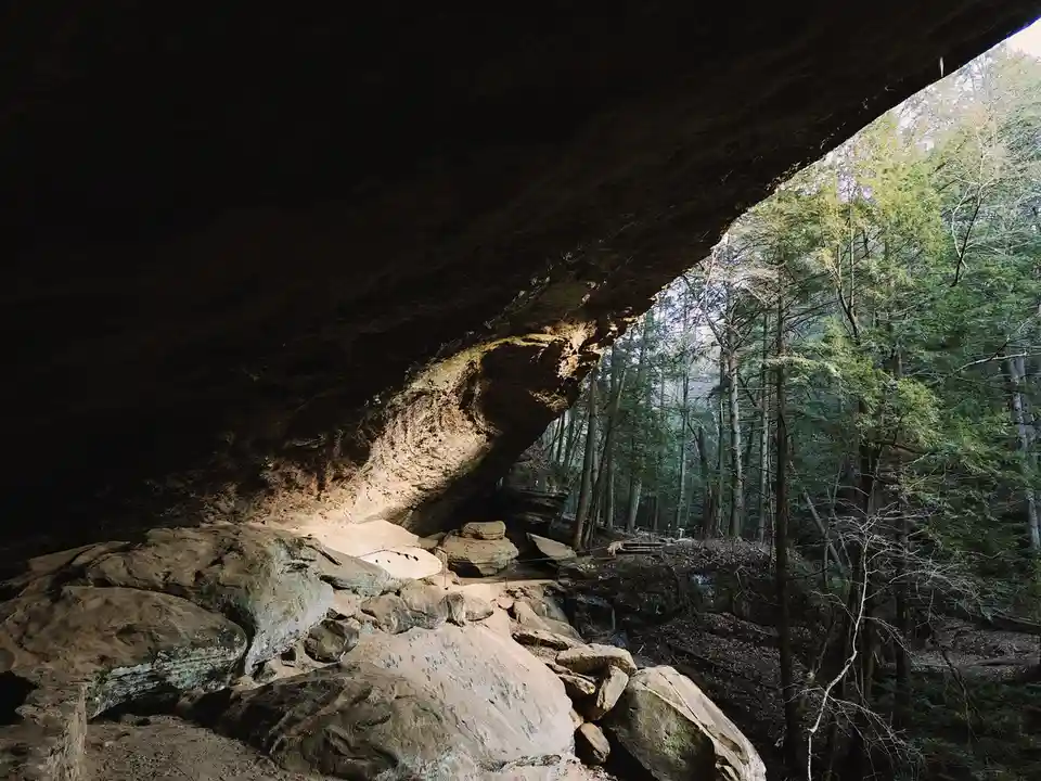 A photo of a large, dark rock overhang inside a forest, with sunlight illuminating the underside of the rock and surrounding…