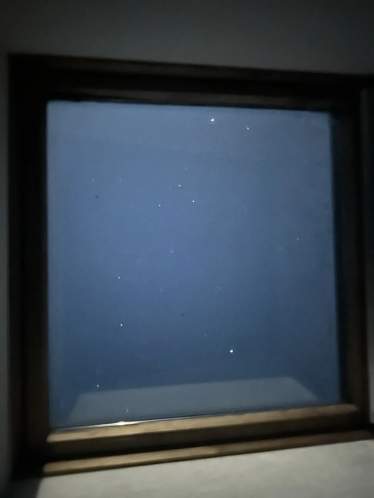 A close-up photo shows a window with a dark wooden frame, looking out onto a night sky filled with faint, scattered stars…