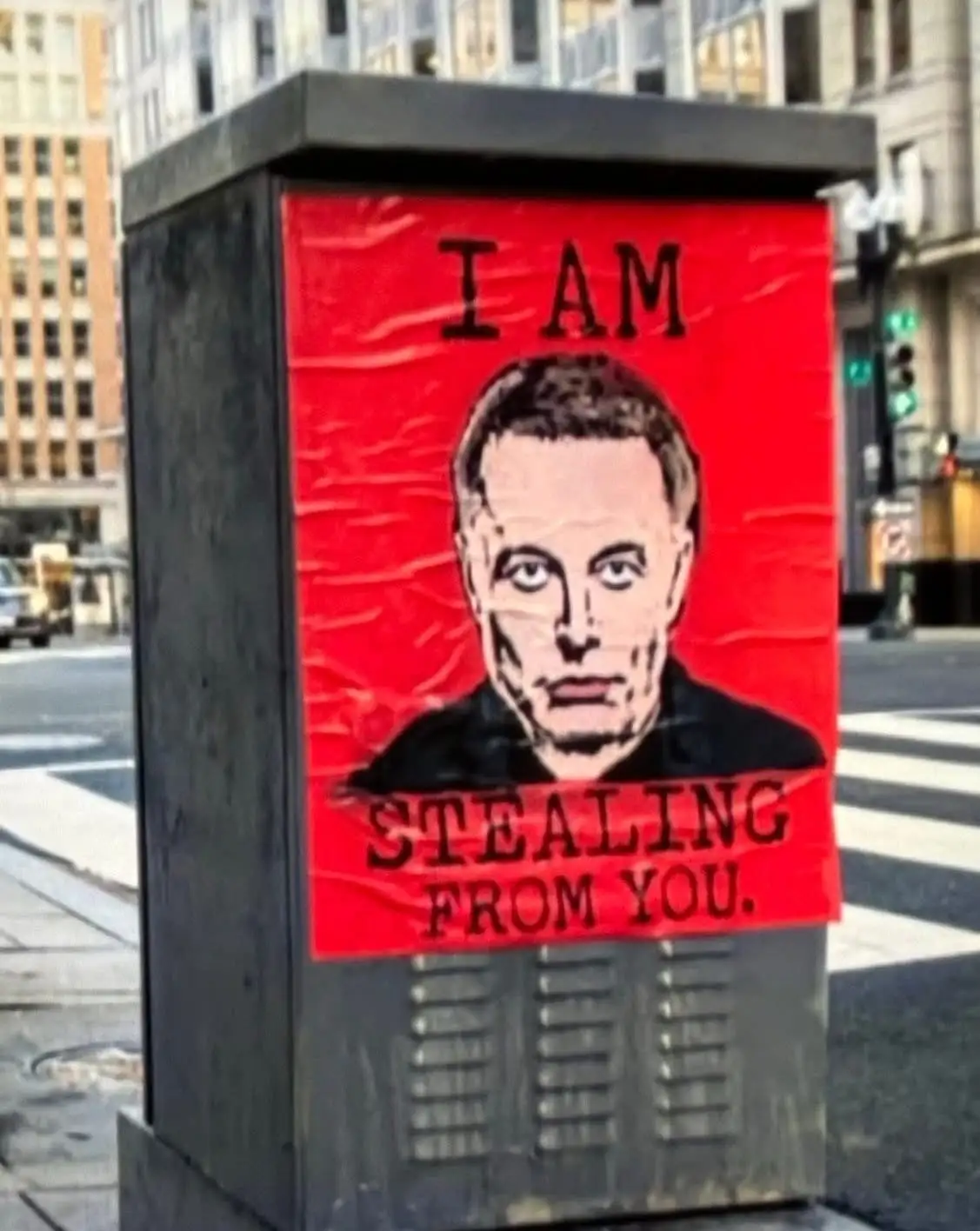 A red poster is taped to a black utility box on a city sidewalk, featuring a stylized portrait of Elon Musk with the bold…