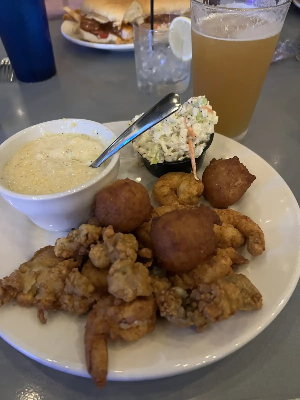 A plate of fried shrimp and hush puppies with a side of creamy sauce and coleslaw, accompanied by a beer and a drink with a…
