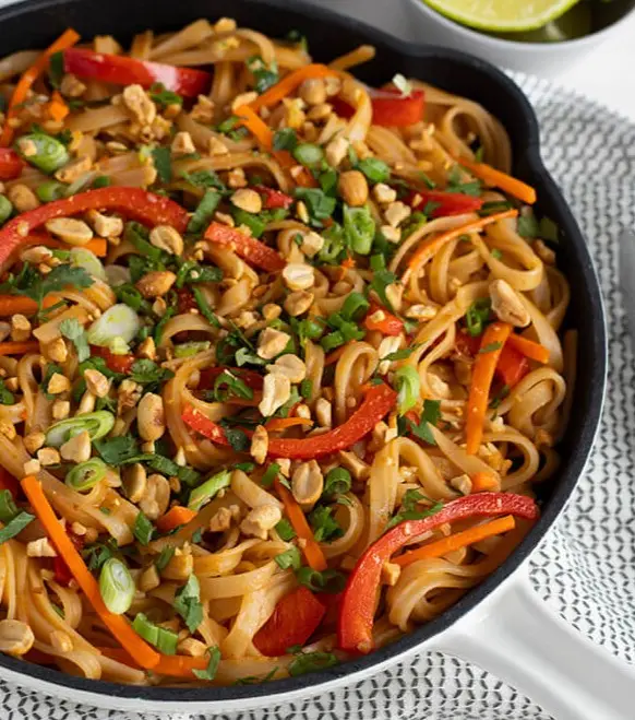 a close-up photo of a skillet filled with pad thai noodles, garnished with chopped peanuts, sliced green onions, and strips…