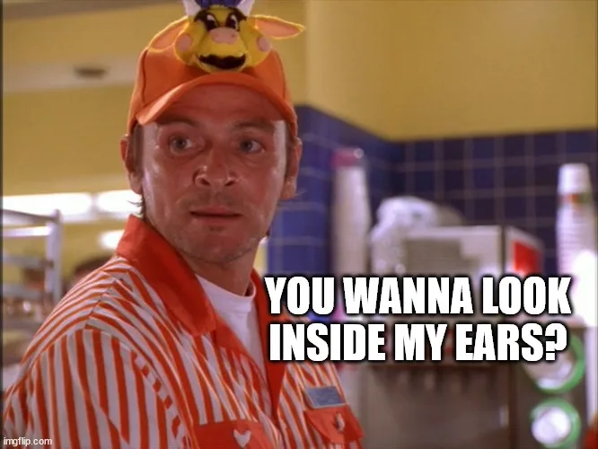 A man in an orange and white striped uniform and an orange cap with a yellow cow plushie on top stares directly at the…