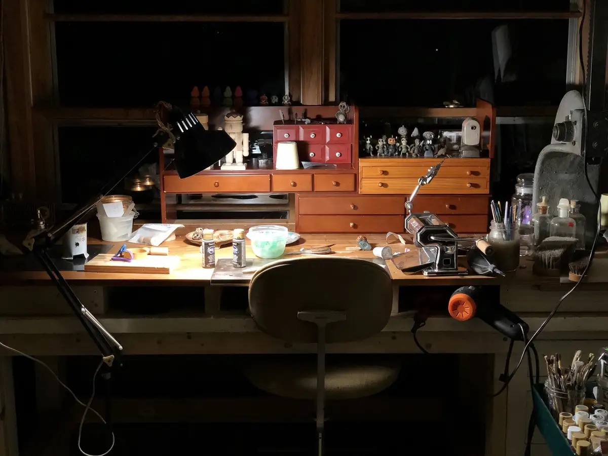 A dimly lit, cluttered wooden workbench at night, illuminated by a single black desk lamp, with various crafting tools…
