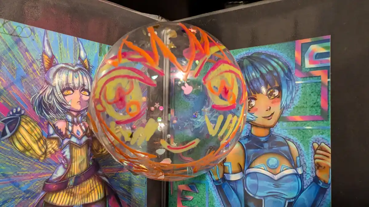 A clear, decorated balloon with colorful scribbles and glitter sits in front of two anime-style posters of female…