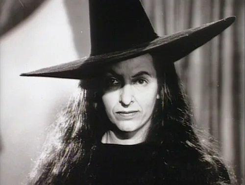 A black-and-white still of a woman with long dark hair wearing a tall, pointed witch's hat, looking directly at the camera…
