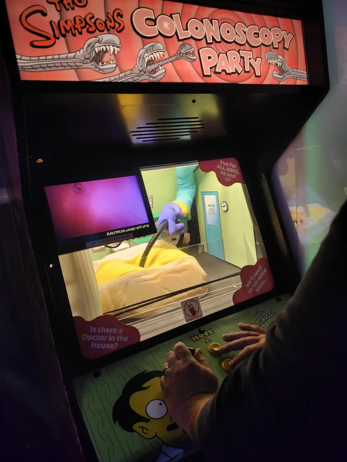 A person plays an arcade game titled 'The Simpsons Colonoscopy Party,' which features a screen showing a colonoscopy…