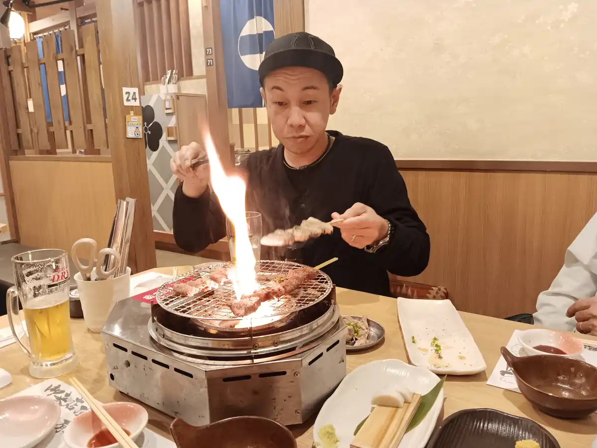 A man in a black cap and shirt is grilling meat at a Japanese restaurant table, holding a skewer over a flaming grill. The…