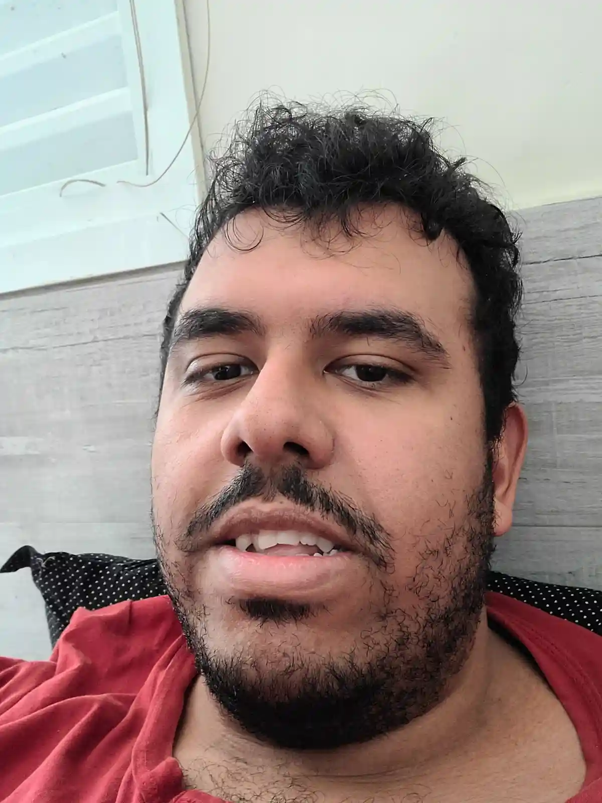 A close-up selfie of a man with dark curly hair, a beard, and mustache, wearing a red shirt, looking directly at the camera…