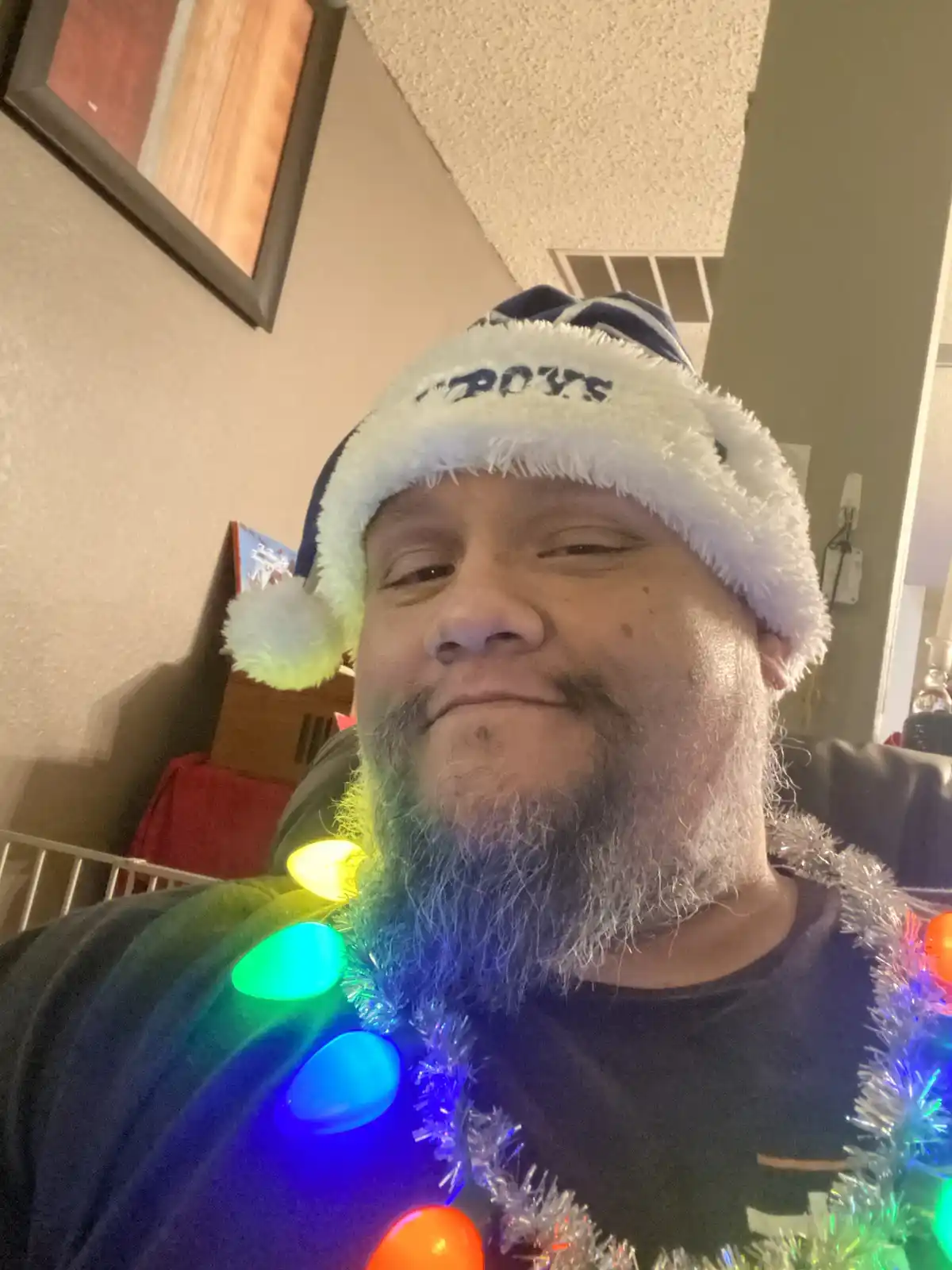 A man with a gray beard wears a blue and white Dallas Cowboys-themed Santa hat and has a string of colorful Christmas lights…