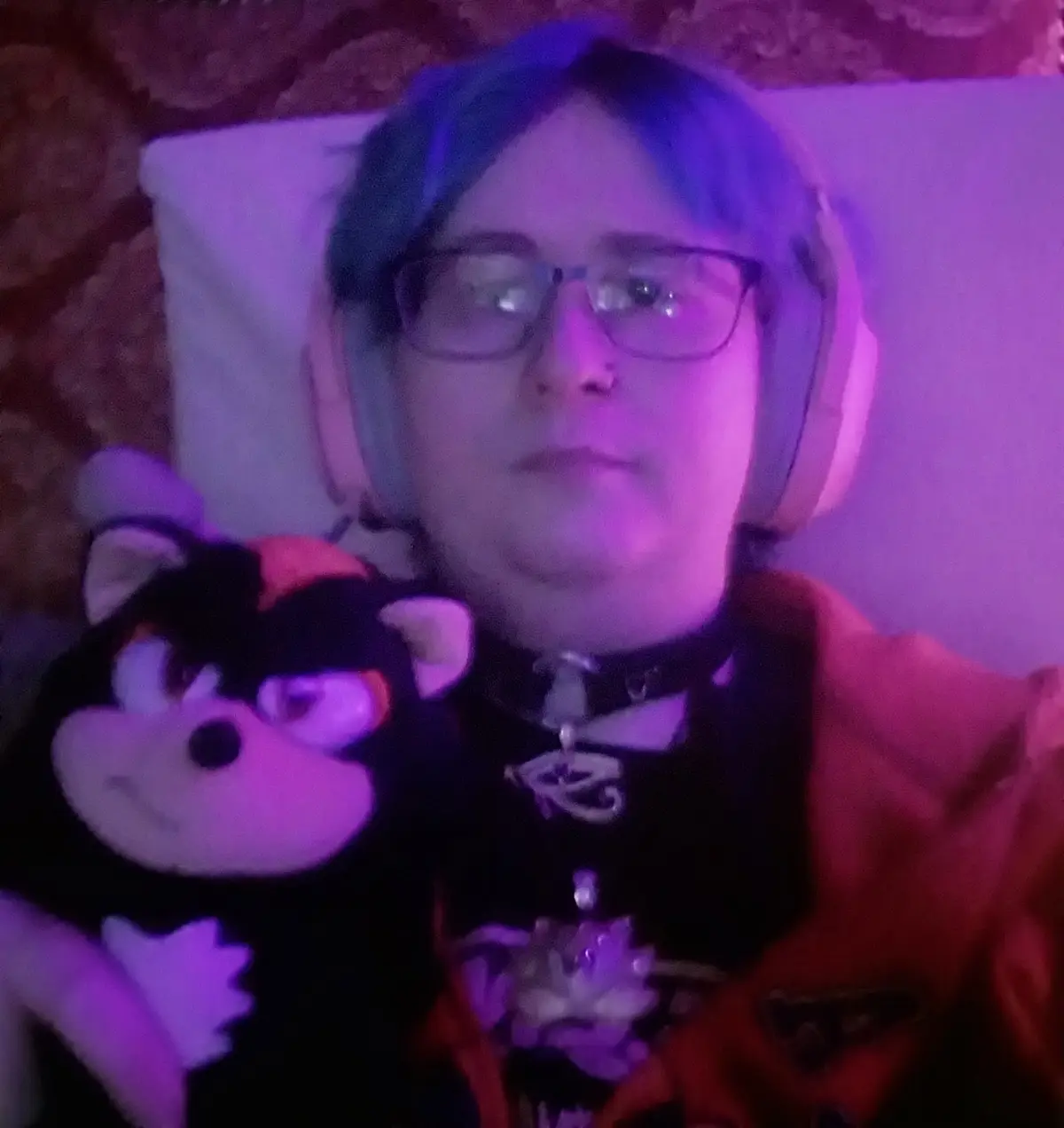 A person with blue hair and glasses, wearing headphones and a red jacket, lies in bed under purple lighting while holding a…