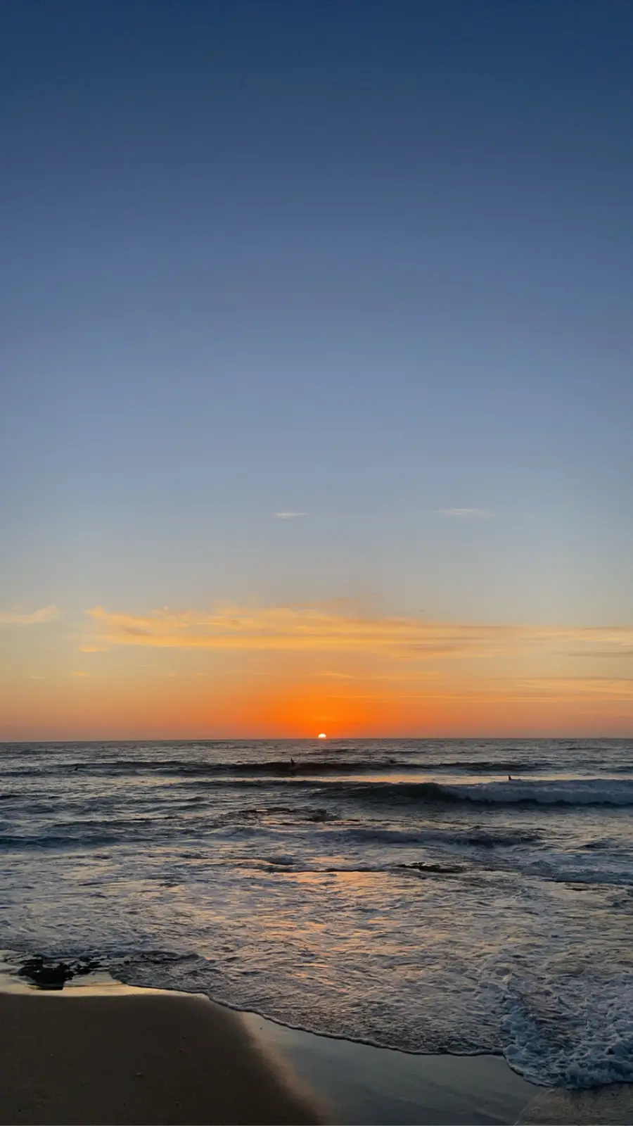 A serene beach sunset photo with the sun low on the horizon, casting orange and blue hues across the sky and water. The…