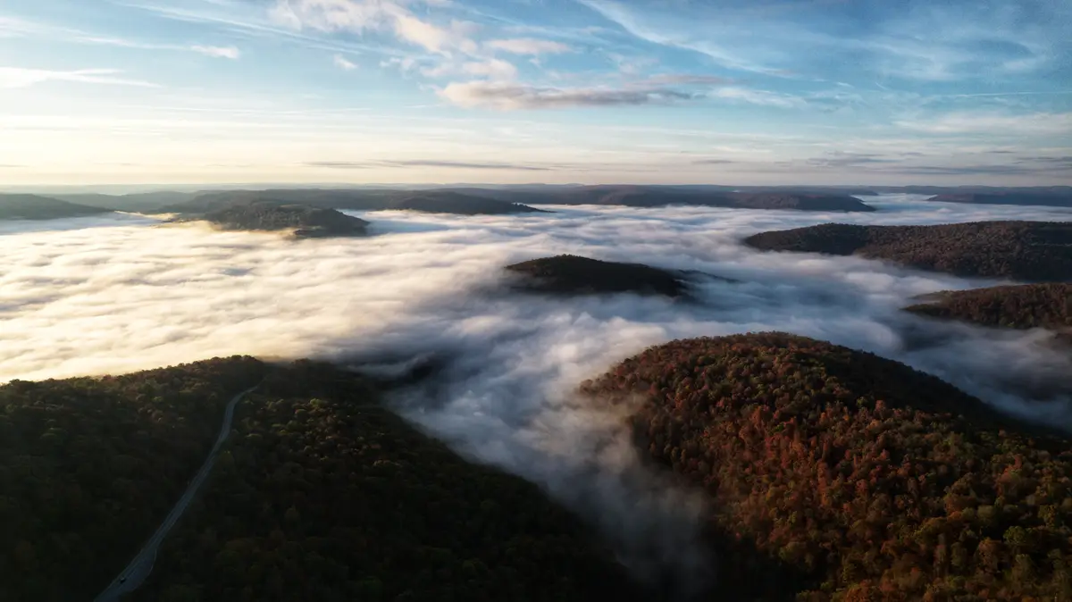 An aerial view of rolling forested hills during sunrise or sunset, with a thick blanket of fog or clouds filling the valleys…