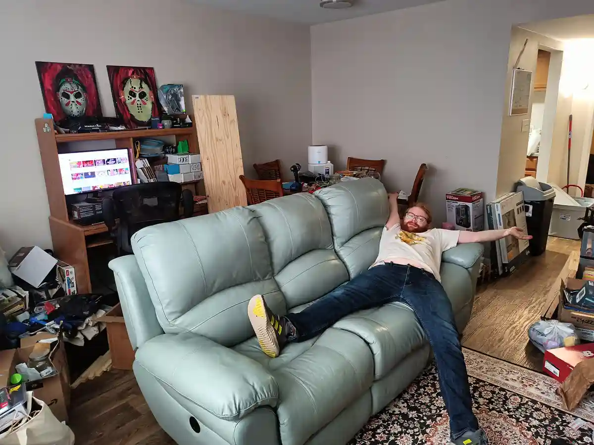 A man with glasses and a beard lies sprawled across a light green leather couch in a cluttered living room, arms…