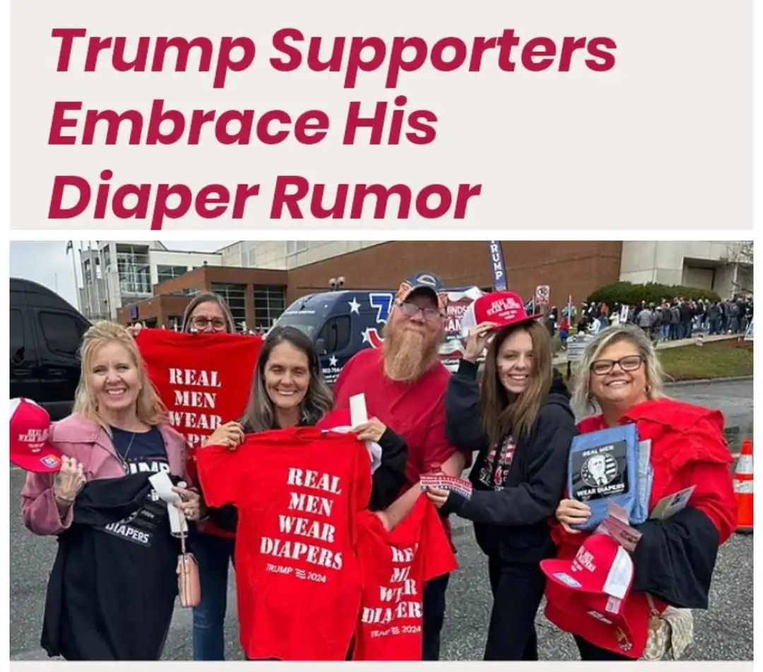 A group of smiling people, mostly women, are posing outdoors holding red t-shirts and hats that say 'REAL MEN WEAR DIAPERS'…