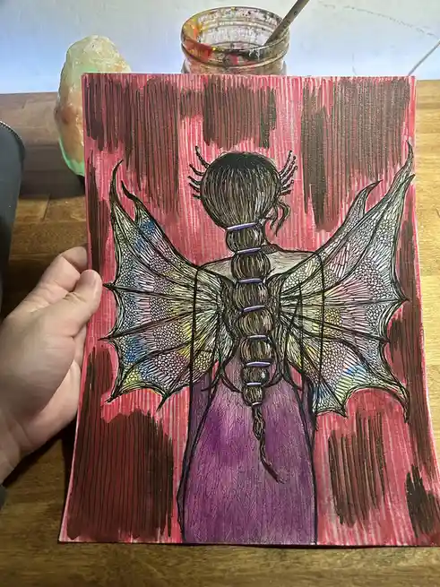 A hand holds up a colorful drawing of a figure seen from behind, featuring large, detailed bat-like wings and a long…