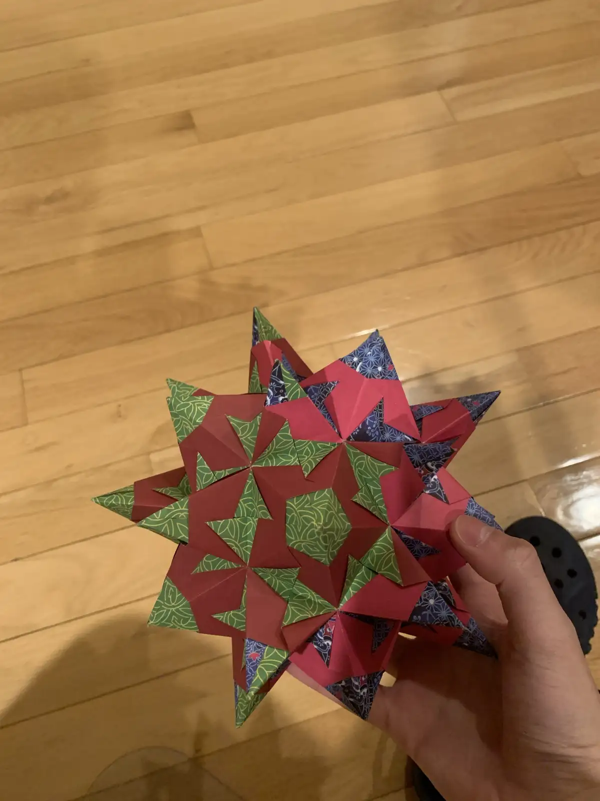 A hand holds a complex, multi-layered origami star made from red, green, and blue patterned paper against a wooden floor…