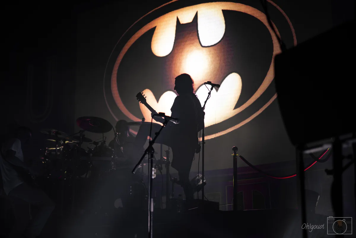 A silhouetted musician plays guitar on a dark stage in front of a large, glowing bat symbol, which is the iconic logo of…
