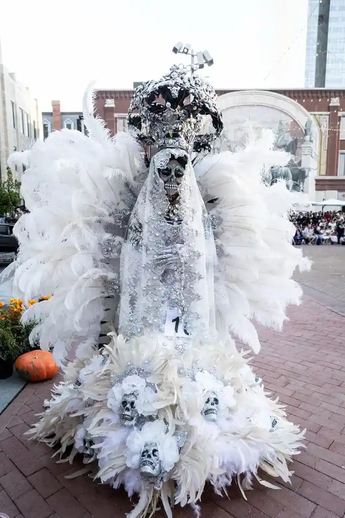 A person in an elaborate, ornate costume resembling a skeletal bride, adorned with white feathers, a jeweled crown, and a…