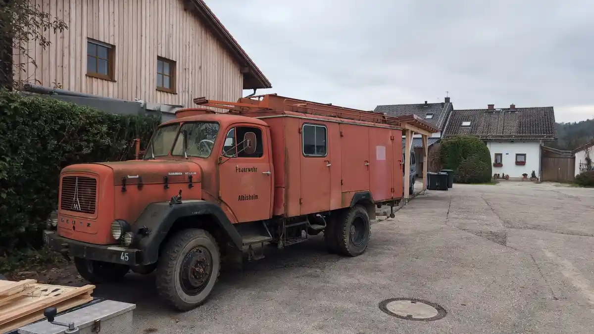 An old, rusty orange Deutz fire truck labeled 'Feuerwehr Albisheim' is parked in a gravel lot beside a wooden building and…