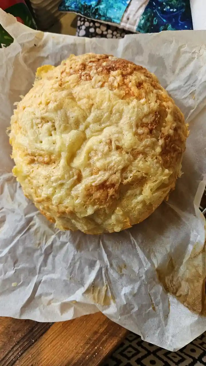 A round, golden-brown baked bread covered in melted, bubbly cheese sits on crumpled parchment paper. The text overlaid on…