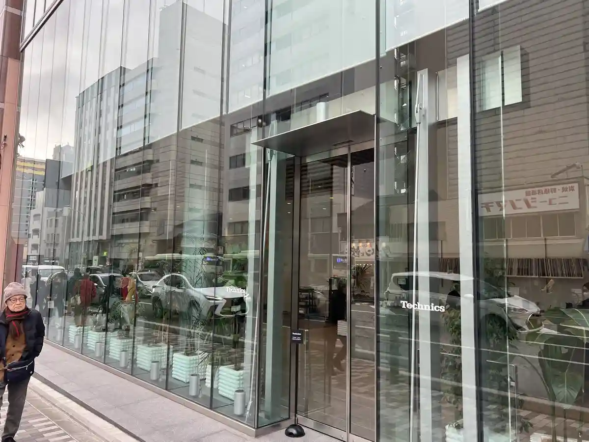 A man in a winter hat and scarf walks past a modern glass-fronted store with 'Technics' written on the doors. Inside, the…