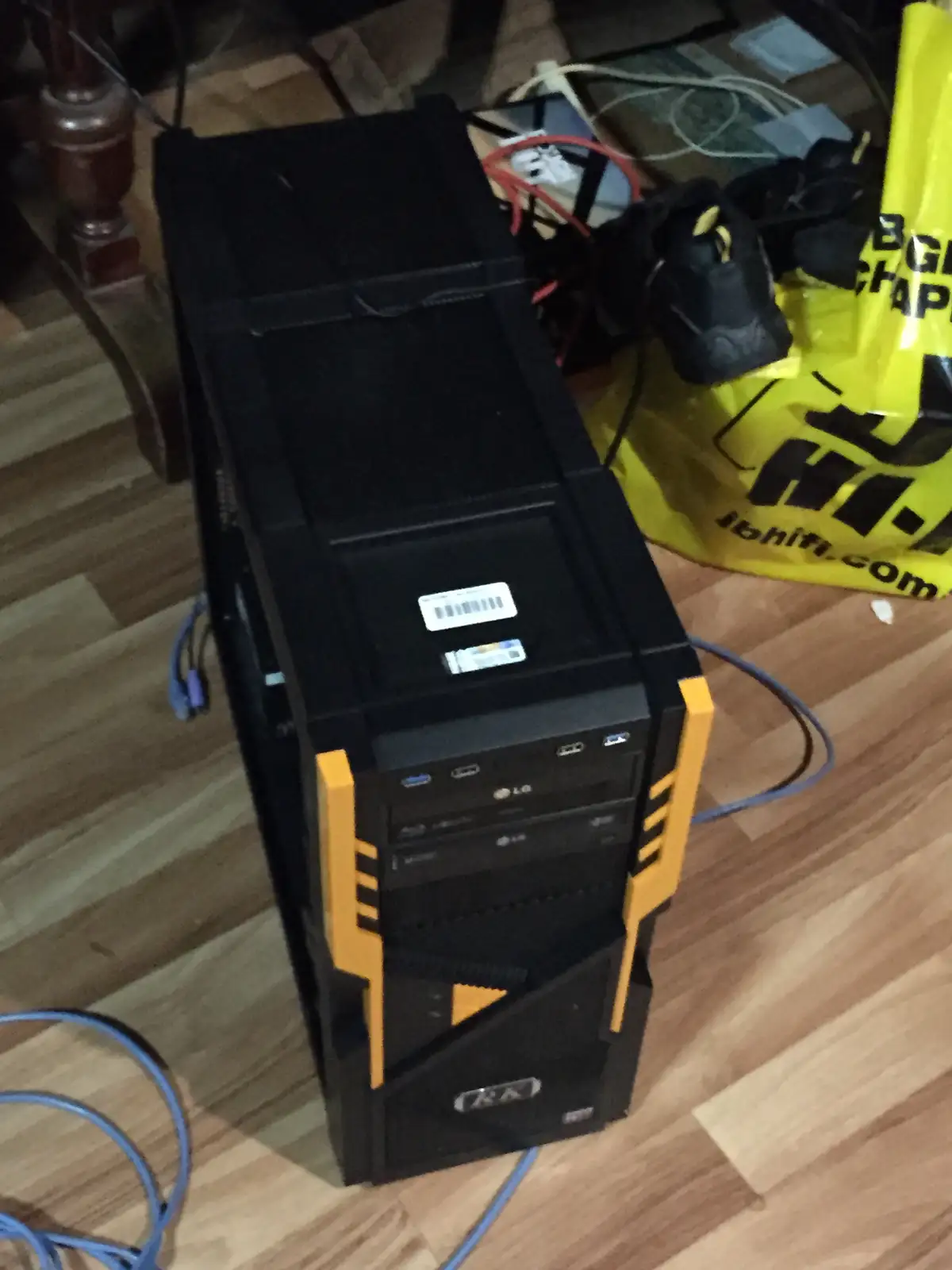 A black and yellow desktop computer tower sits on a wooden floor next to a yellow 'ibhifi.com' bag and tangled cables. The…