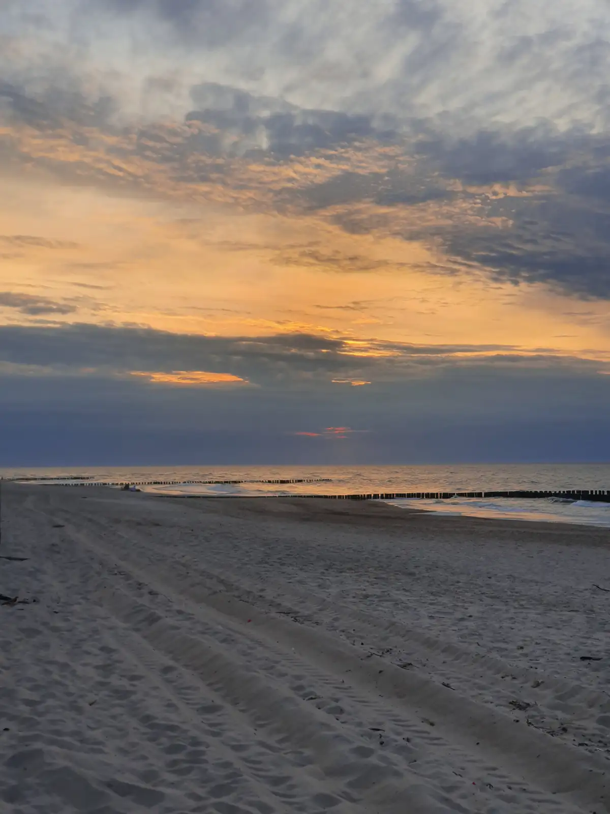 A serene beach scene at sunset with orange and gray clouds, calm ocean waves, and tire tracks in the sand. The overlaid text…