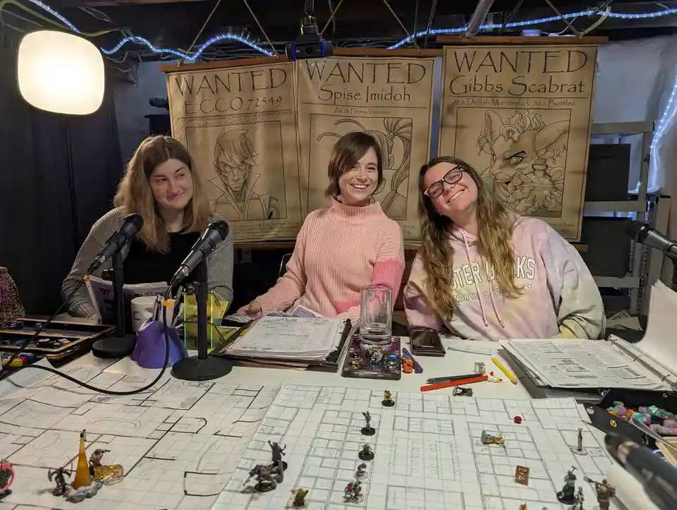 Three women sit at a table with microphones and miniature figurines on grid paper, surrounded by 'WANTED' posters featuring…