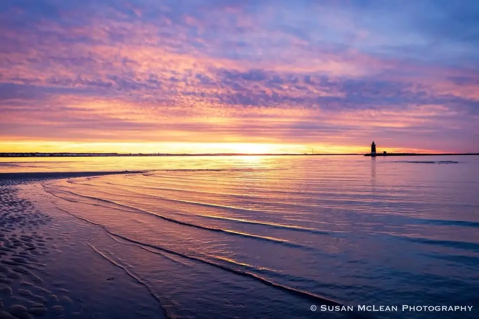 A serene sunset photograph shows gentle waves rolling onto a sandy beach under a vibrant sky of purple, pink, and orange…
