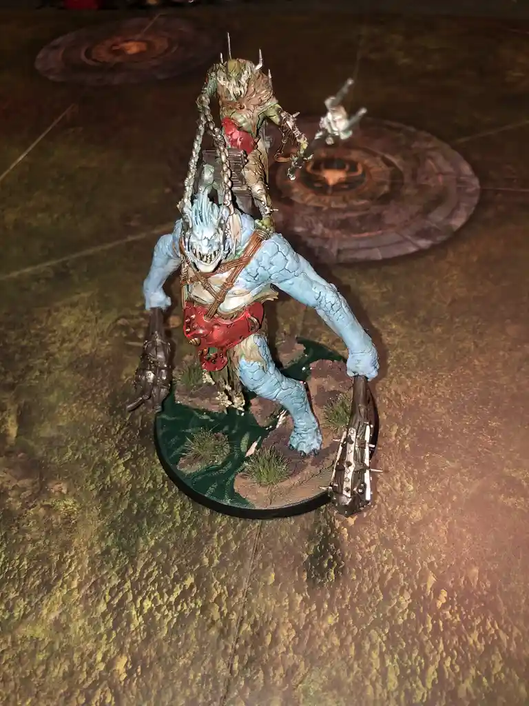 A painted miniature of a blue-skinned, monstrous creature with a spiked helmet and chains, standing on a circular base with…