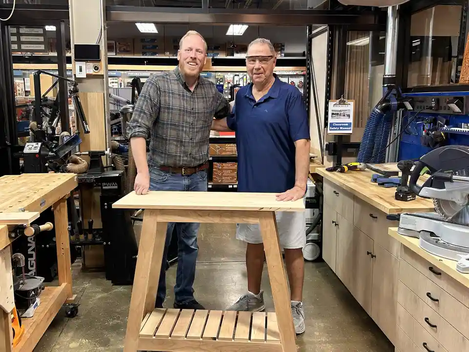 Two men stand in a woodworking workshop behind a small wooden table they built, smiling at the camera. The younger man on…