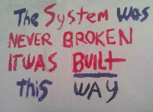 A handwritten sign in red and blue marker on white paper reads 'The System Was NEVER BROKEN IT WAS BUILT- this WAY', with…