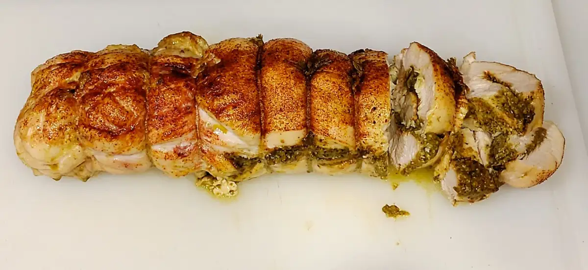 a roasted, sliced chicken breast with visible green pesto stuffing sits on a white cutting board. The text overlaid on the…