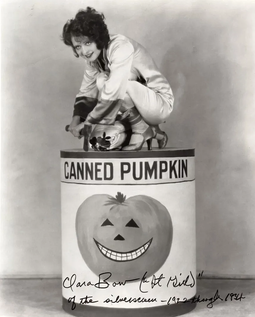 A vintage black-and-white photo shows actress Clara Bow crouching atop a giant can labeled 'CANNED PUMPKIN', which features…
