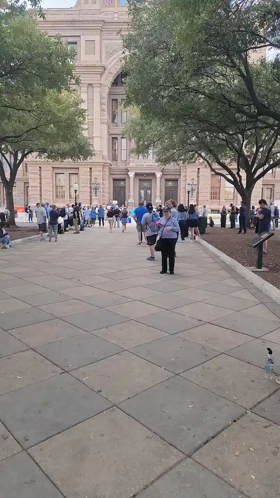 A wide-angle photo shows a large crowd of people gathered on a paved plaza in front of a grand, classical-style stone…