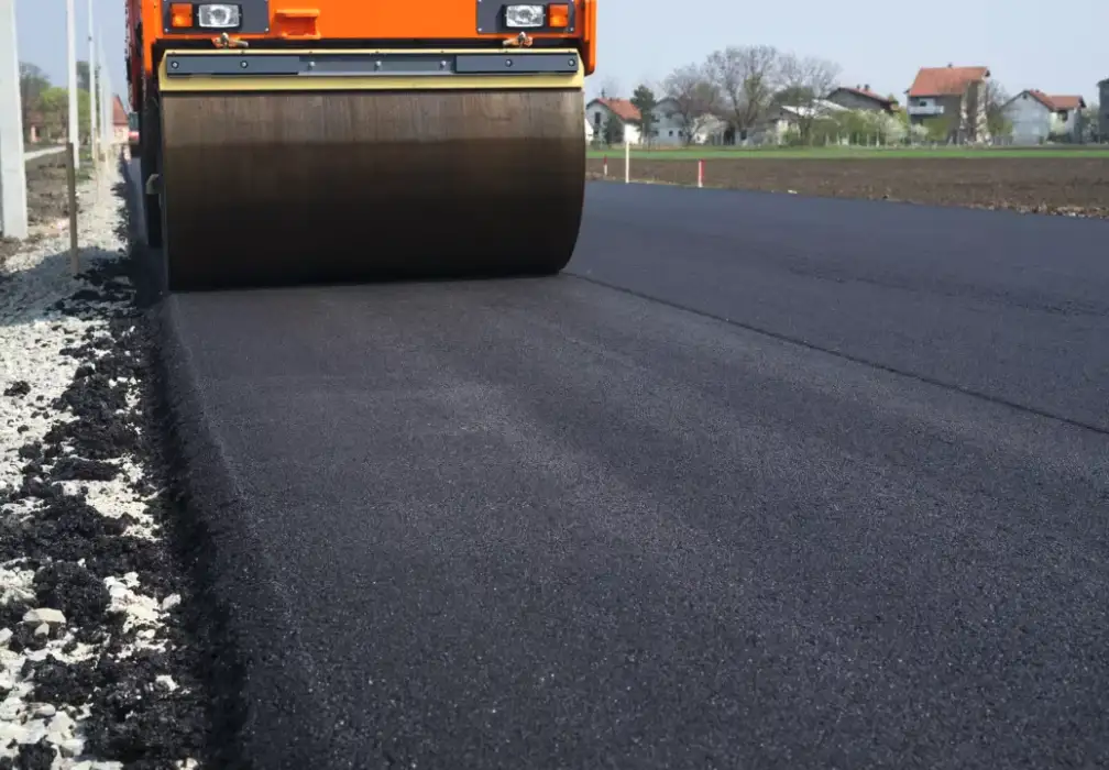 A bright orange road roller is compacting fresh black asphalt on a new road, with a rough gravel edge and residential houses…