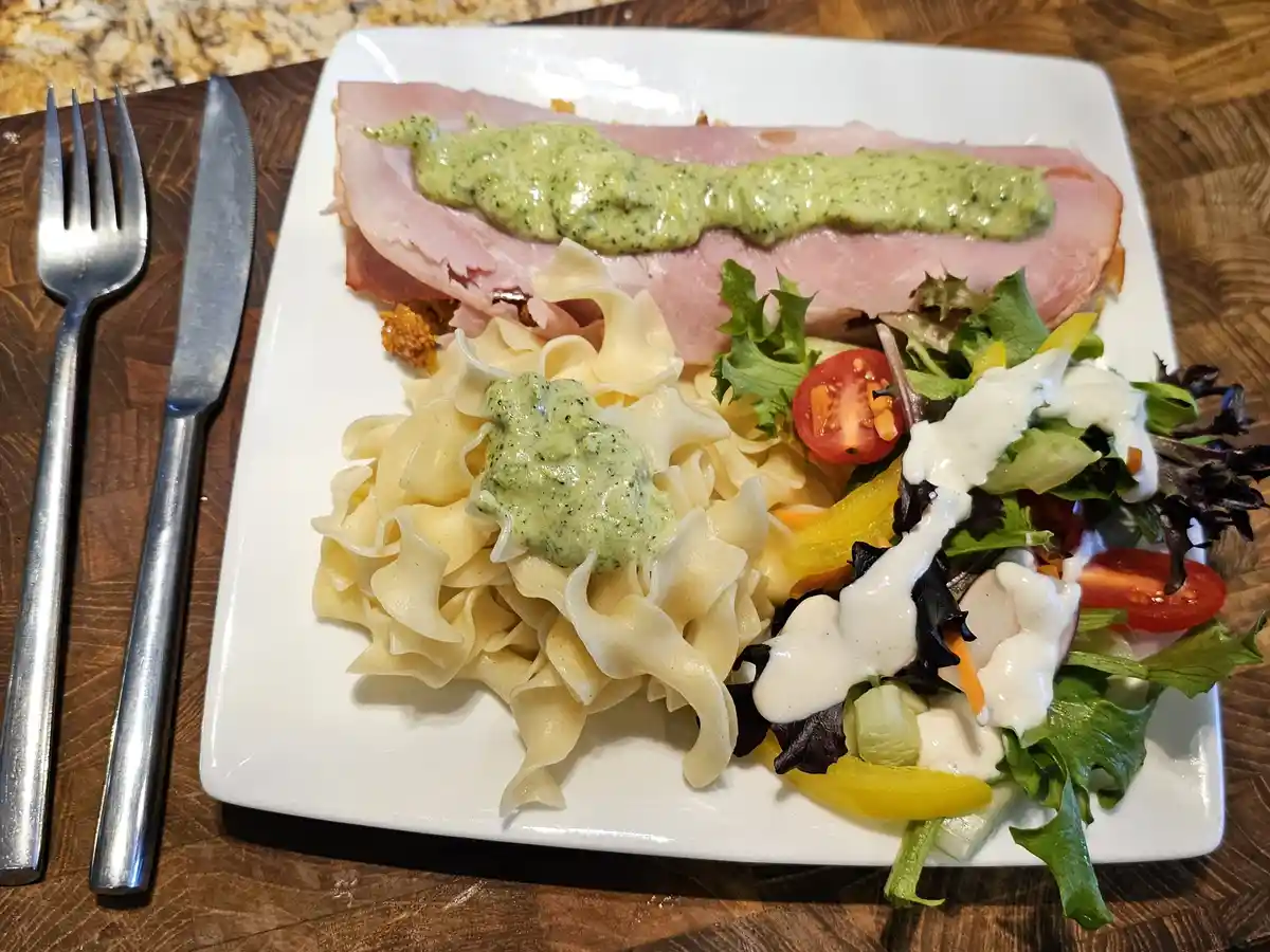 A white plate holds a meal with ham, pasta, and salad, all topped with green sauce and white dressing. A fork and knife rest…