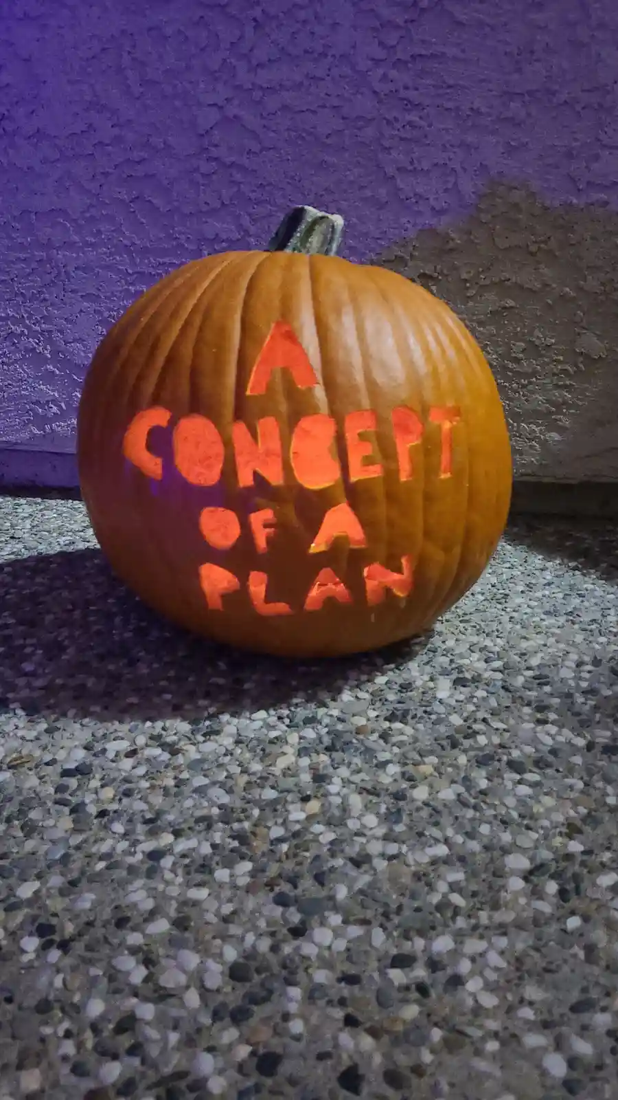 A carved orange pumpkin with the text 'A CONCEPT OF A PLAN' illuminated from within, sitting on a pebbled concrete surface…