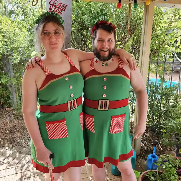 Two people stand arm-in-arm outdoors wearing identical green and red elf-themed knit dresses with candy cane buttons and…