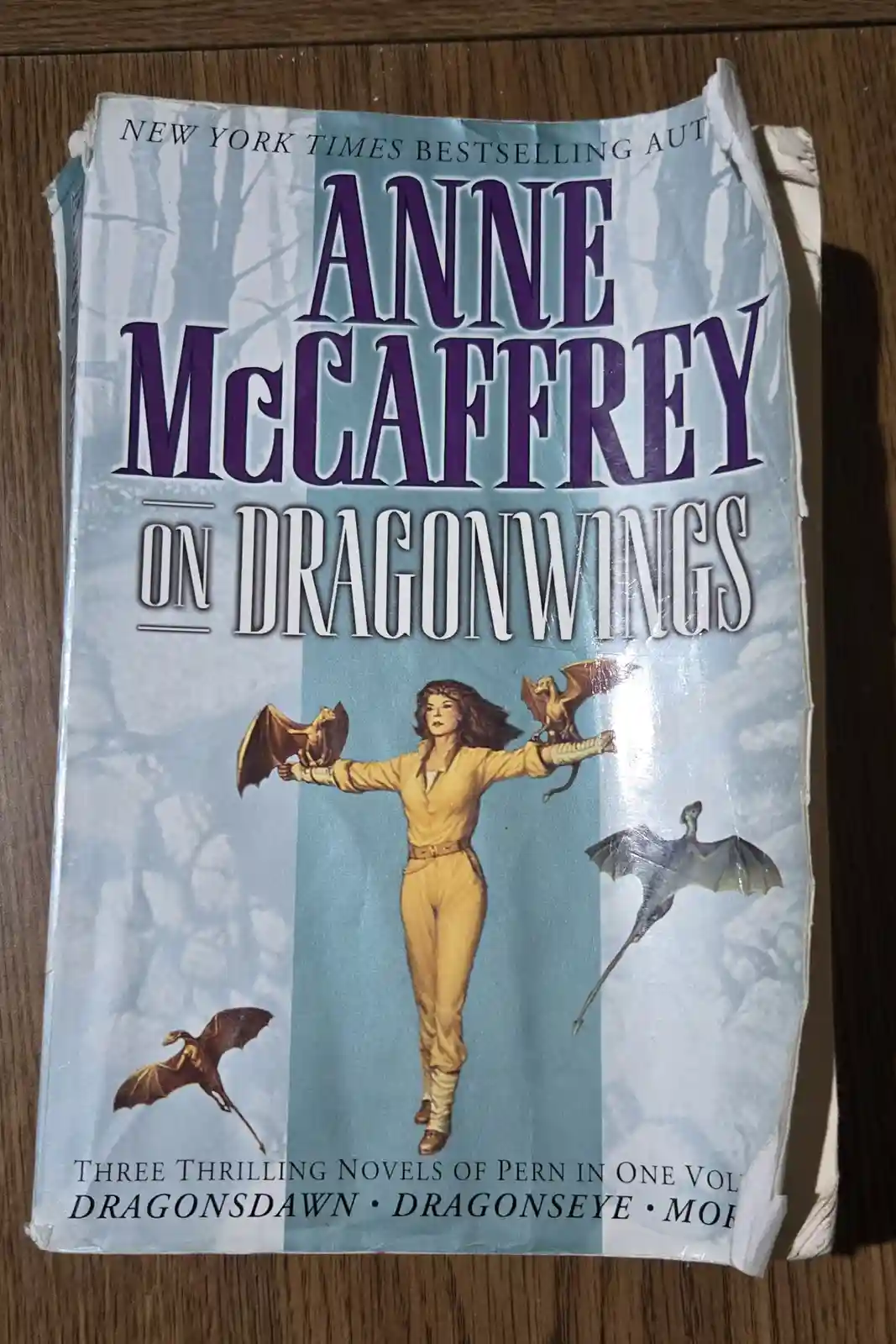 A worn paperback book cover for 'On Dragonwings' by Anne McCaffrey, showing a woman in yellow flight gear with dragons on…