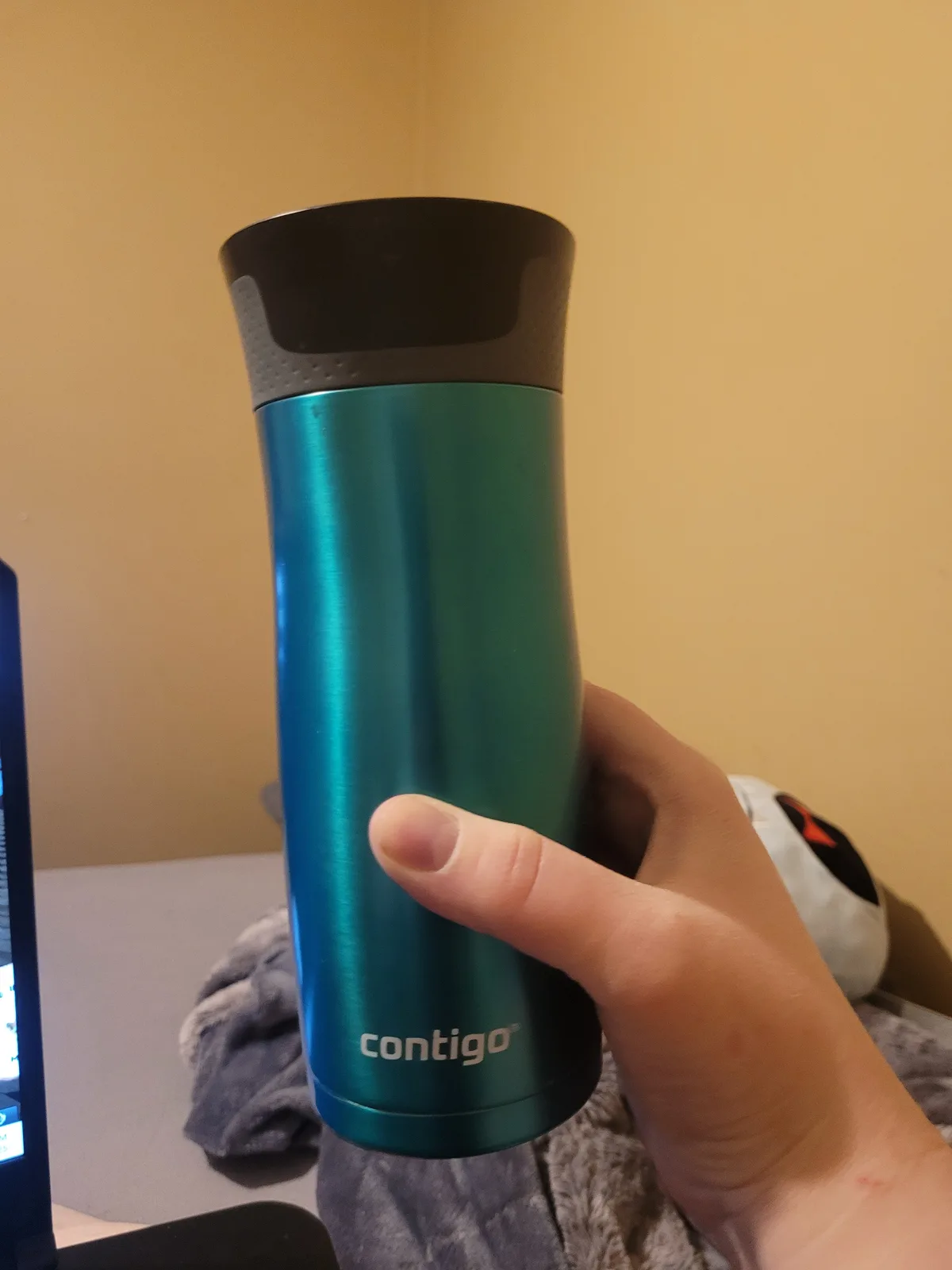 A hand holds a teal-colored Contigo brand travel mug with a black lid against a beige wall, with a laptop and plush toy…
