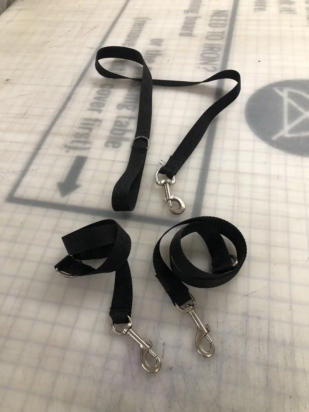 Three black nylon dog leashes with metal clasps lie on a white gridded ironing board cover that has text like 'NEED TO…