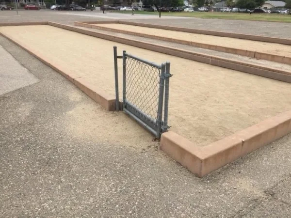 The image shows an outdoor sand pit with raised concrete borders, featuring a small chain-link gate that is open and leaning…