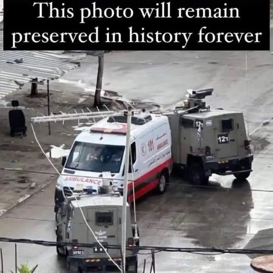 An aerial photo shows a white ambulance with red stripes and Arabic/English text parked next to two grey armored military…
