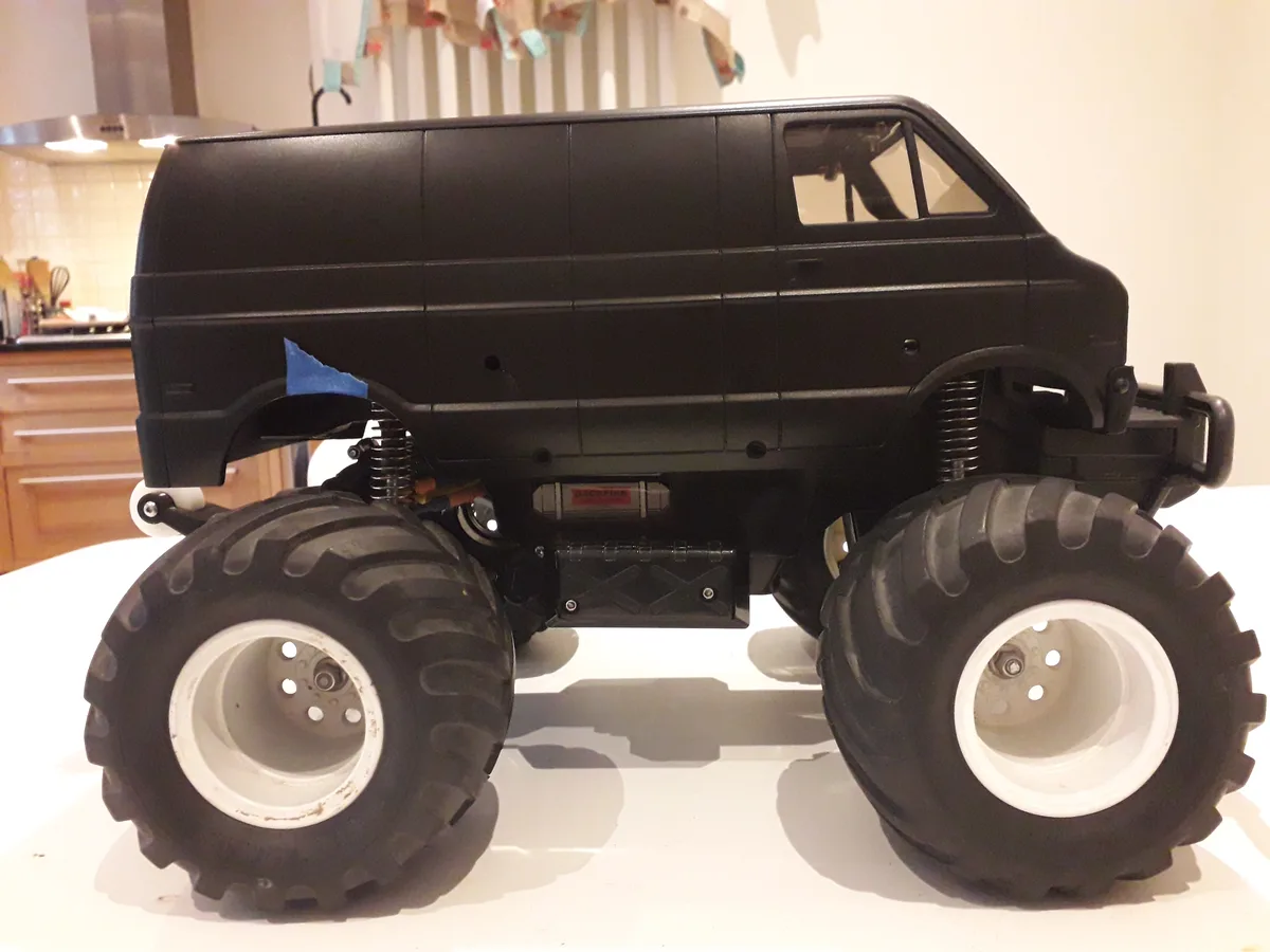 A black remote-controlled monster truck van with oversized black tires and white rims sits on a white kitchen counter. The…