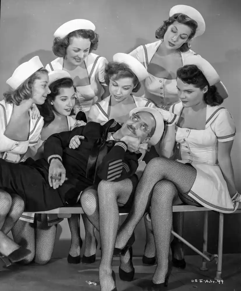A vintage black-and-white photo shows a man in a sailor uniform reclining on a bench, surrounded by six women in sailor…