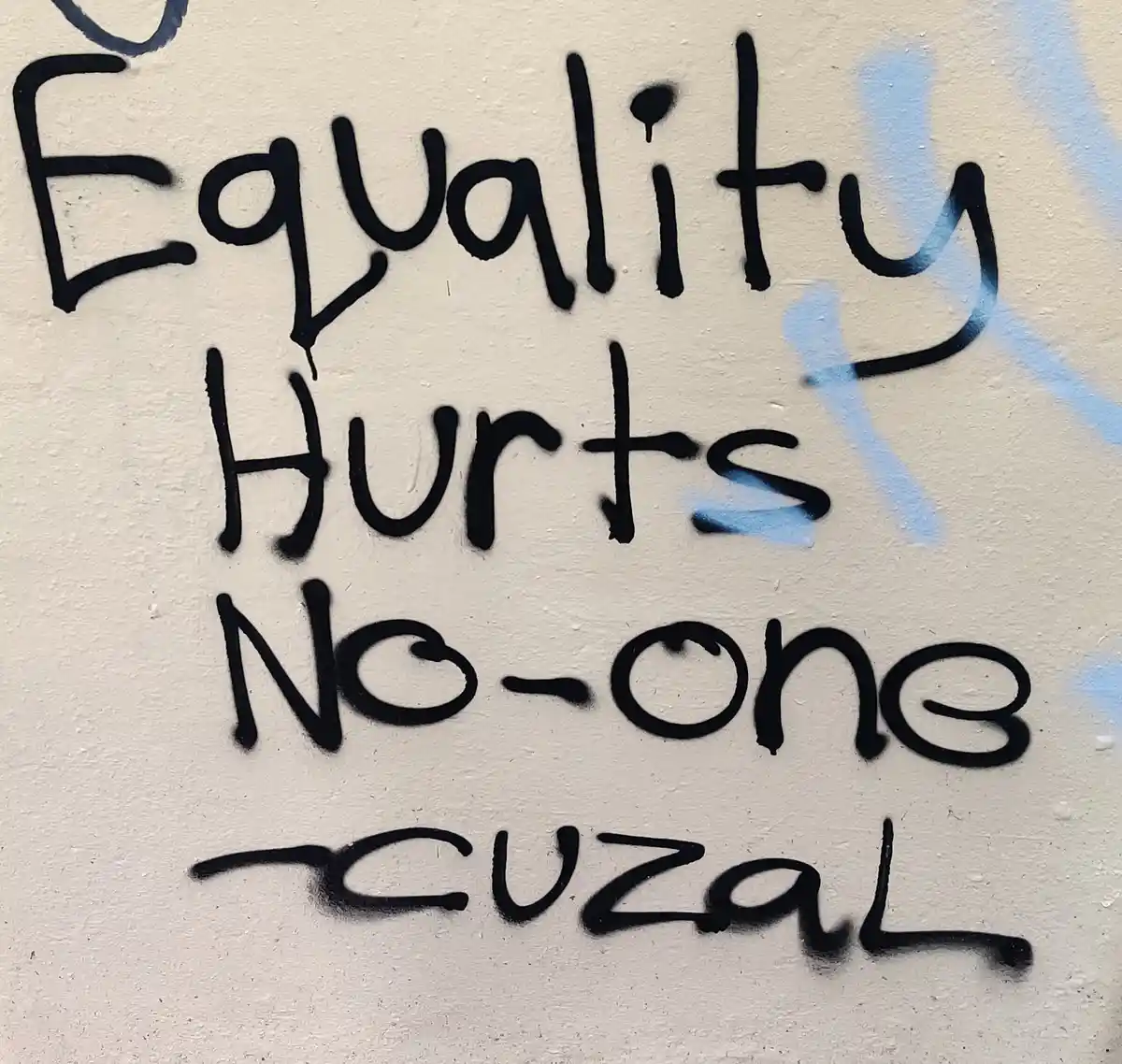 A close-up of black spray-painted text on a light-colored wall, reading 'Equality Hurts No-one cuzal' in a casual, slightly…