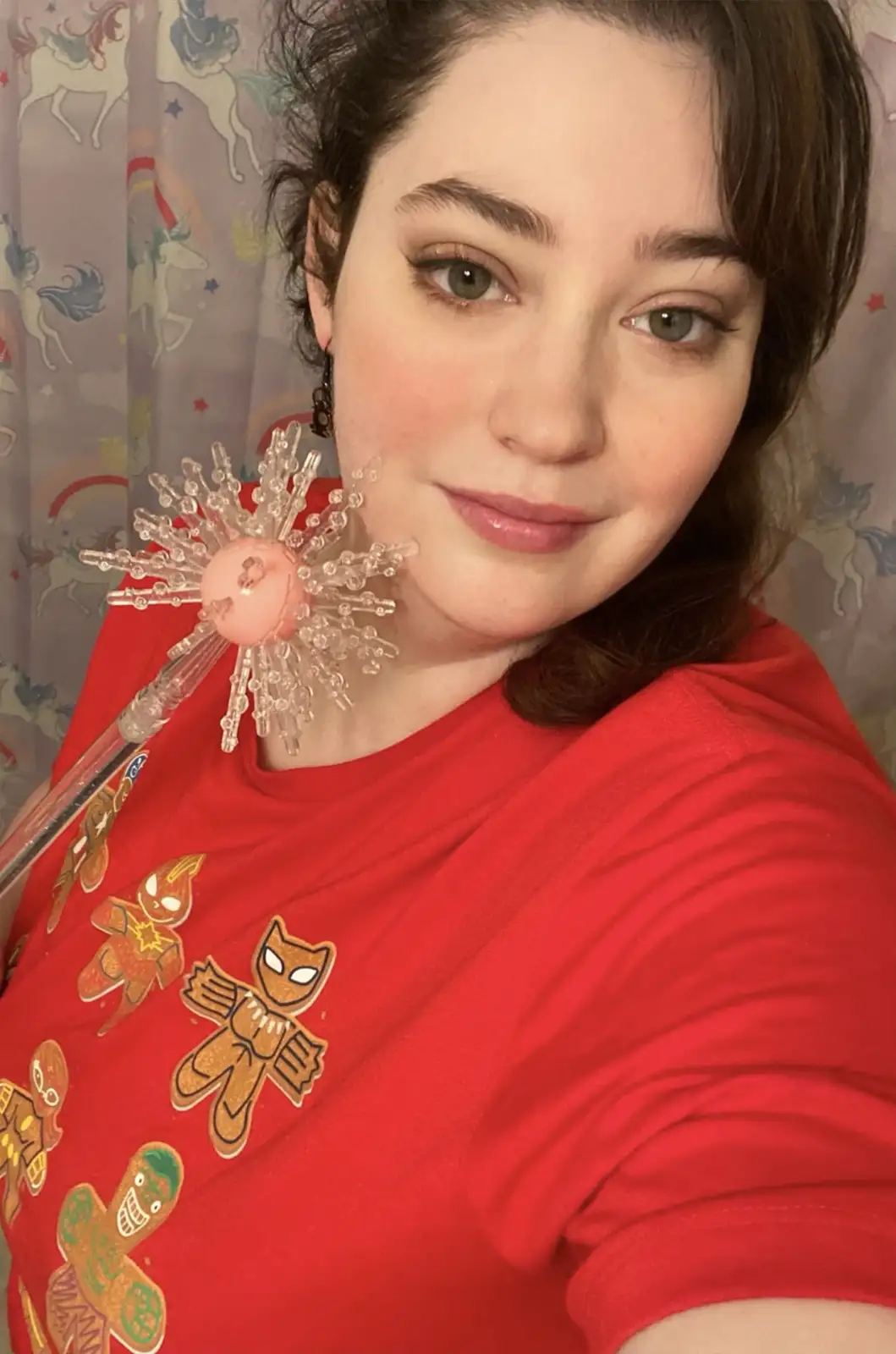 A woman with brown hair and green eyes wears a red t-shirt with cartoon gingerbread men on it and holds a large, clear…